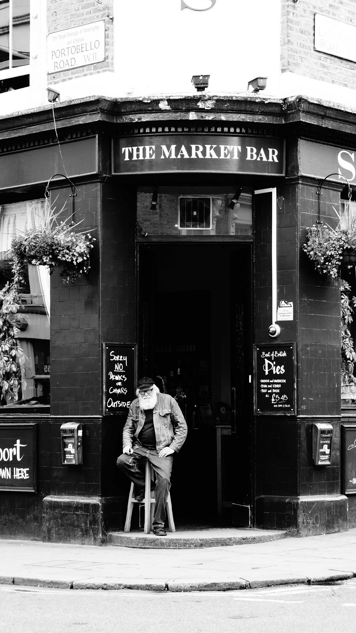 An old man sitting on a bar chair in front of 'The Market Bar'. Just sitting there as if part of the scene..