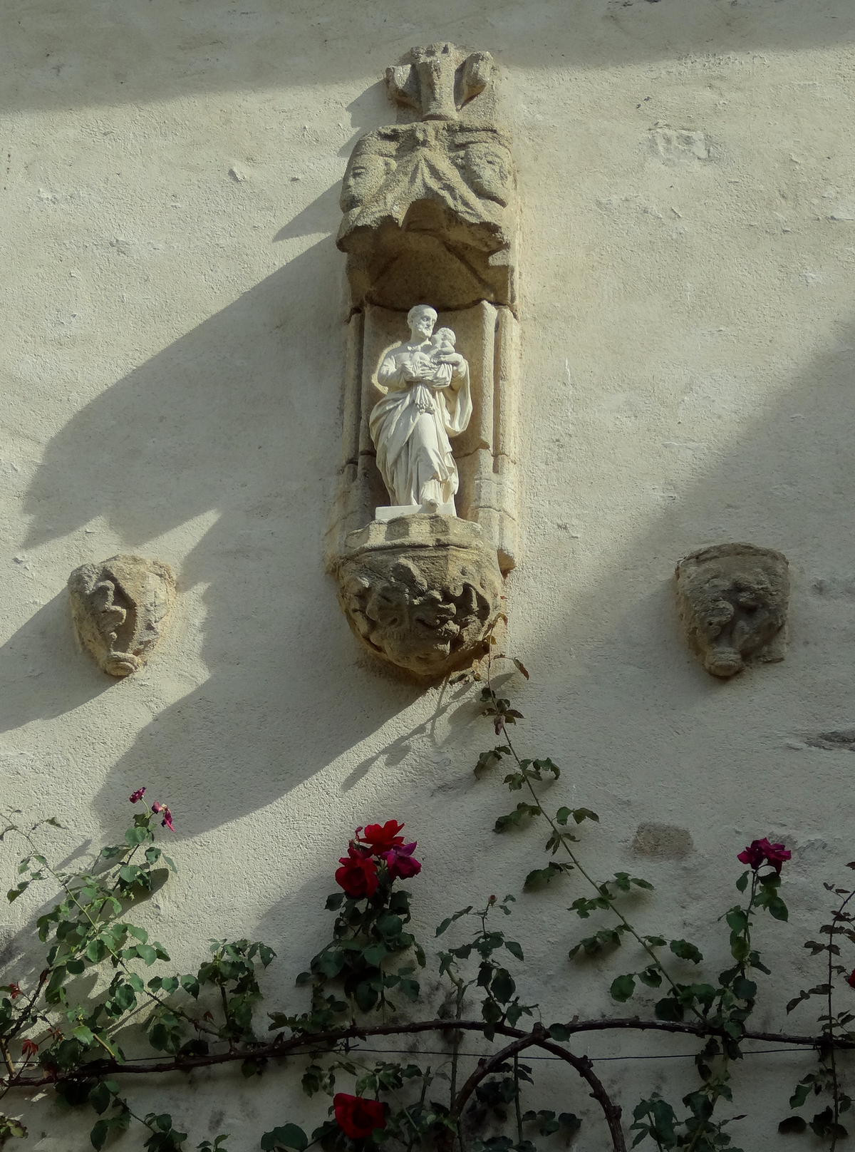 Dans la cour d'un ancien prieuré, Saint Joseph tenant dans ses bras l'enfant Jésus. La lumière oblique du soleil déclinant, un rosier aux fleurs rouges dans l'ombre des murs.